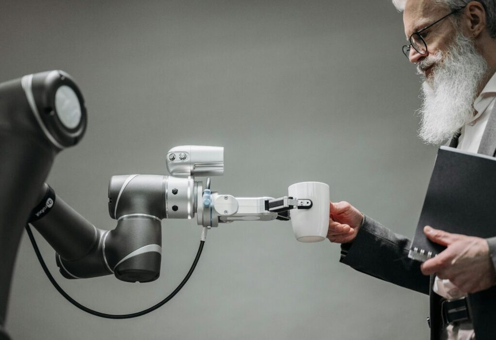 An elderly man receives a cup from a robotic arm in a modern office setting. &mdash; Photo by Pavel Danilyuk on Pexels